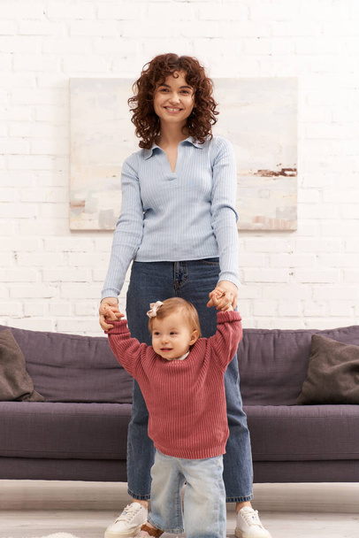 first steps, quality family time, bonding, balancing work and life, happy working mother holding hands with toddler daughter, togetherness, cozy living room, denim jeans, casual attire - Photo, Image