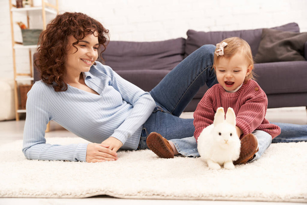 engaging with kid, happy and curly woman sitting on carpet with toddler daughter in cozy living room, playing with rabbit, quality family time, casual attire, bonding between mother and child  - Photo, Image