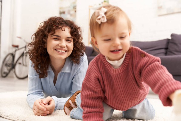 engaging with kid, happy and curly woman lying on carpet with toddler daughter in cozy living room, quality family time, casual attire, bonding between mother and child  - Photo, Image