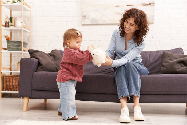 engaging with kid, happy and curly woman sitting on couch with toddler daughter in cozy living room, playing with rabbit, quality family time, casual attire, bonding between mother and child  - Foto, imagen