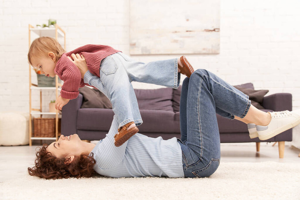 качественное семейное время, счастливая мать поднимая малыша дочь и лежа на ковре в уютной гостиной, баланс трудовой жизни, джинсовая одежда, случайный наряд, семейные отношения, современное воспитание  - Фото, изображение