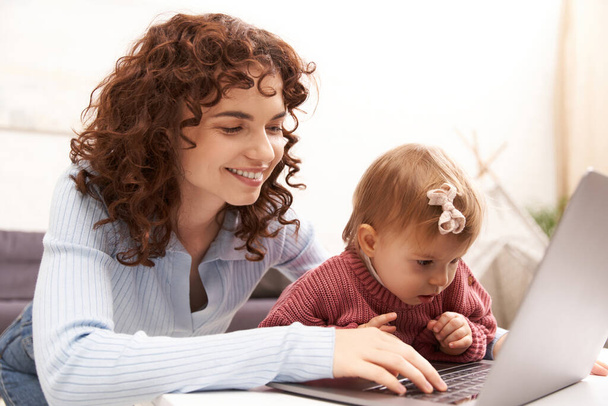 multitasking woman, freelance, modern working parent, balancing work and life, cheerful woman using laptop in cozy living room, modern parenting, building successful career, engaging with child  - Foto, immagini