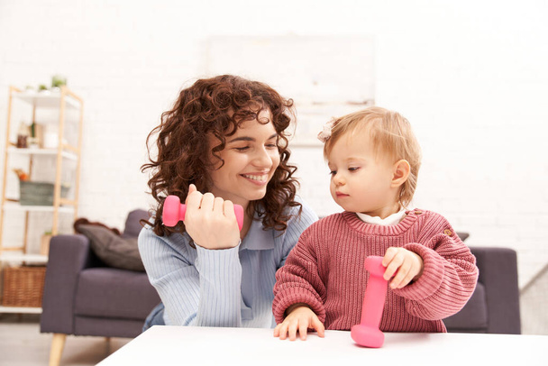 time management, working mother, balanced lifestyle, curly woman exercising with dumbbell near toddler daughter in cozy living room, home workout, sport, busy mom, modern parenting  - Photo, Image
