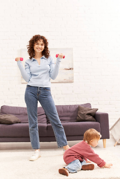 time management, working mother, balanced lifestyle, curly woman exercising with dumbbells near toddler daughter in cozy living room, home workout, sport, busy mom, physical activity  - Photo, Image