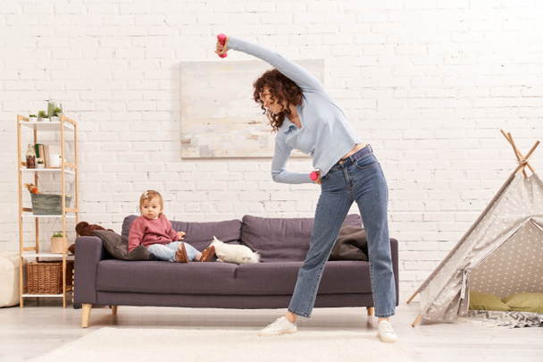 busy mom, modern parenting, working mother, balanced lifestyle, curly woman exercising with dumbbells near toddler daughter on couch in cozy living room, home workout, sport, time management  - Photo, Image