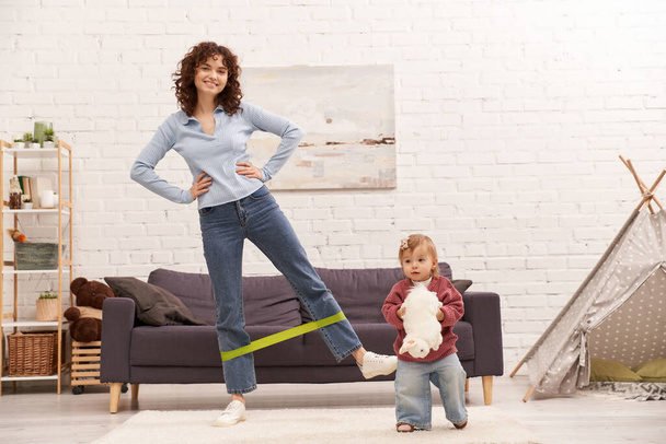 busy mom, modern parenting, working mother, balanced lifestyle, happy woman exercising with resistance band near toddler daughter with soft toy in cozy living room, home workout, sport  - Foto, imagen