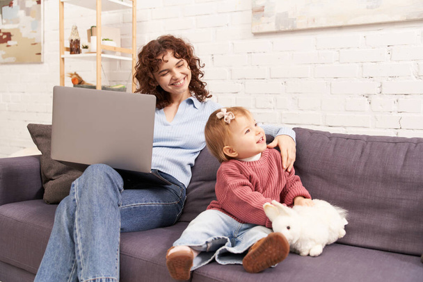 multitasking woman, freelance, curly woman using laptop and sitting on couch with baby girl in cozy living room, modern parenting, building successful career, balancing work and life  - Фото, зображення