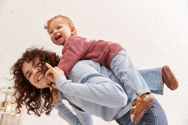 quality time, happiness, balancing between work and life, cheerful woman with excited baby girl on back, mom daughter time, having fun together, bonding, loving motherhood, curly hair  - Foto, Bild