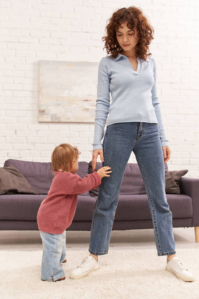 full length of toddler baby doing her first steps near curly mother, cozy living room, engaging with kid, denim jeans, casual attire, family time, modern parenting, work life balance  - Photo, Image