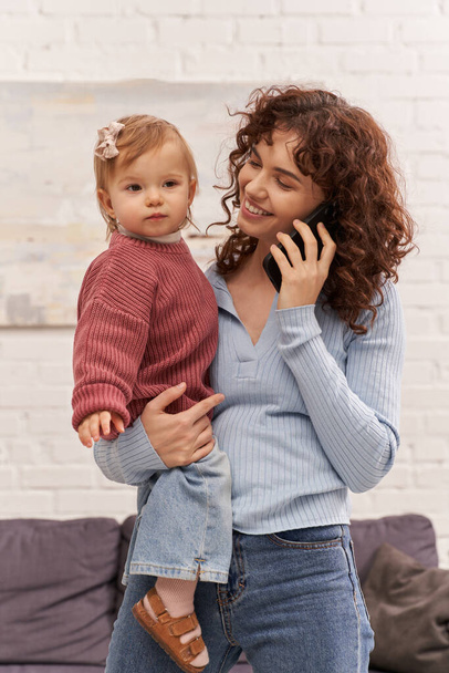 happy young woman holding in arms baby girl and talking on smartphone, balancing between work and life, modern parenting, relationships, mother daughter bond, remote work, career and family  - Fotografie, Obrázek