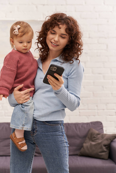 kudrnatá mladá žena drží v náručí dítě a pomocí smartphonu, rovnováha mezi prací a životem, moderní rodičovství, vztahy, matka dcera pouto, vzdálená práce, kariéra a rodina  - Fotografie, Obrázek