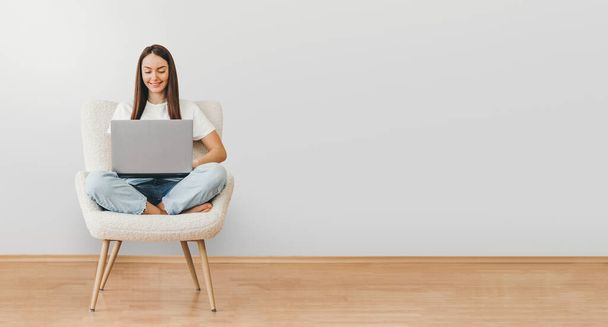 portrait of caucasian woman holding laptop computer while sitting on a chair isolated over gray background. web banner. copy space - Фото, изображение