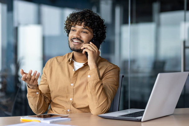 Young successful man working inside office on laptop at workplace, businessman in shirt talking cheerfully smiling on phone, hispanic man with curly hair and casual shirt. - Zdjęcie, obraz