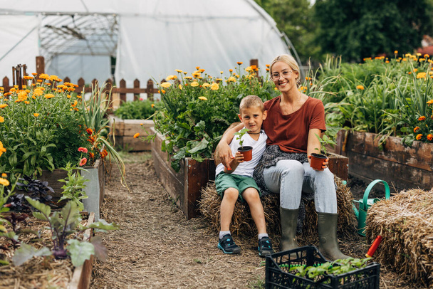 A boy s sitting on a haystack with his mom in their sustainable garden. - Foto, Imagen