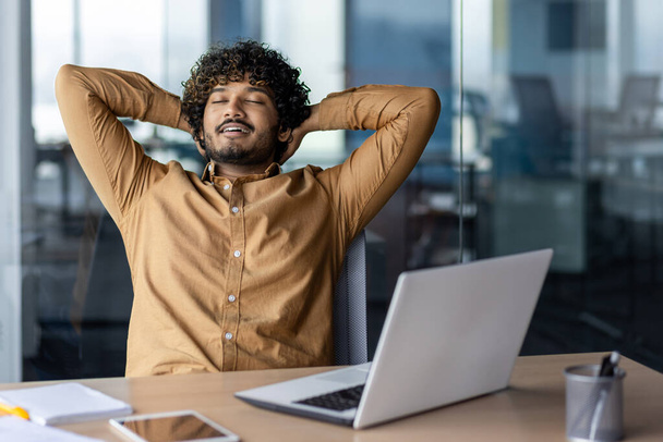 Task successfully completed and job well done, businessman typing away inside office at workplace, satisfied hispanic man with hands behind head dreaming and smiling with closed eyes. - Foto, Bild