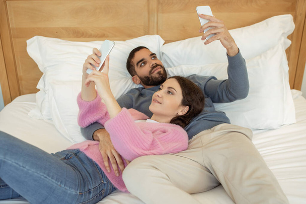 Mobil Teknoloji ve İnsanlar. Genç evli çift internette sörf yapıyor, sosyal medyada mesajlaşıyor ve evde yatakta uzanırken eğleniyor. Modern Gadgets Yaşam Tarzı. Seçici Odaklanma - Fotoğraf, Görsel