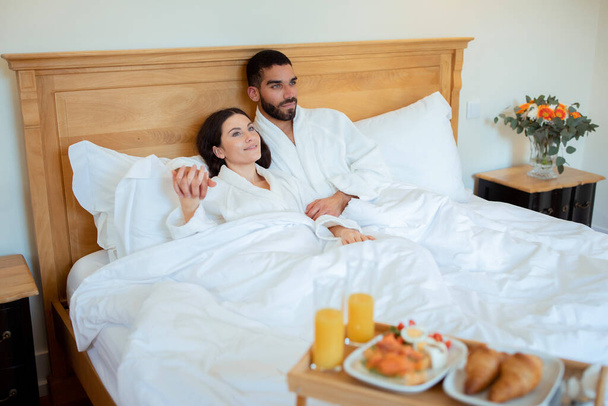 Утром медового месяца. Пара влюбленных обнимаются в удобной кровати, с подносом для завтрака для них утром в отеле Интерьер. Супруги отдыхают в праздничное утро. Роскошь и отдых - Фото, изображение