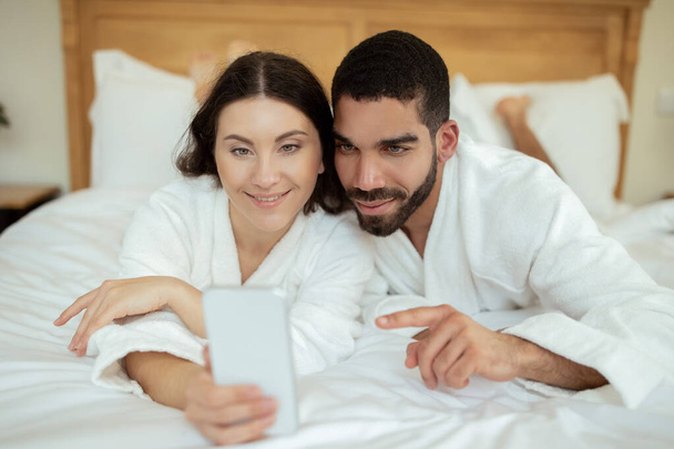 モバイルアプリ。ベッドに横たわっスマートフォンを使用して陽気なカップル,朝の居心地の良いモダンなホテルのインテリアで白いバスローブを身に着けているポーズ,テキストとオンライン通信.ファミリーガジェット - 写真・画像