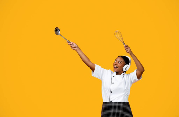 Happy Black Chef Femme dans les écouteurs dansant sur fond jaune studio, joyeux afro-américain cuisinière dame tenant des ustensiles et pointant du doigt à côté de l'espace de copie, faire bouger Dab, s'amuser - Photo, image