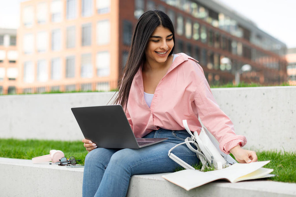 ワークブックやノートパソコンで勉強する笑顔の学生女性、公園や大学のキャンパスで屋外に座っている間に本を読んでコンピュータを使用する女性、フリースペース - 写真・画像