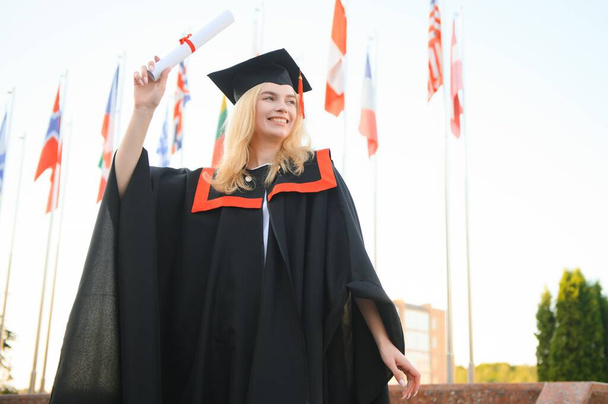 Portrait enthusiastic female college student graduate in cap and gown celebrating, holding diploma - 写真・画像
