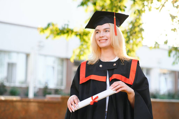 Happy cute caucasian grad girl is smiling. She is in a black mortar board, with red tassel, in gown, with nice brown curly hair, diploma in hand. - 写真・画像