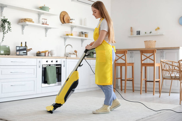 Young woman vacuuming carpet in kitchen - Valokuva, kuva