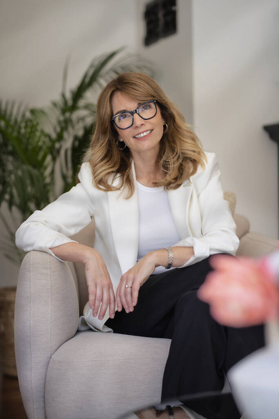 Happy attractive woman relaxing in an armchair at home. Blond haired female wearing eyeglasses and white blazer. - Foto, imagen