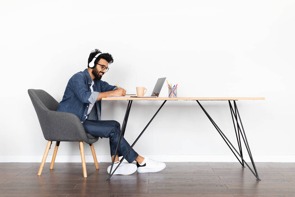 Cheerful handsome young middle eastern guy sit at desk, using laptop, wireless headphones, taking notes, millennial man attend online training, webinar from home, white wall background, copy space - Foto, Imagen