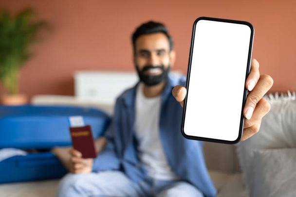 Новое приложение для путешествий. Счастливый арабик показывает большой экран смартфона и паспорт с билетами на самолет дома. Молодой турист показывает отличное мобильное приложение для путешественников, макет - Фото, изображение