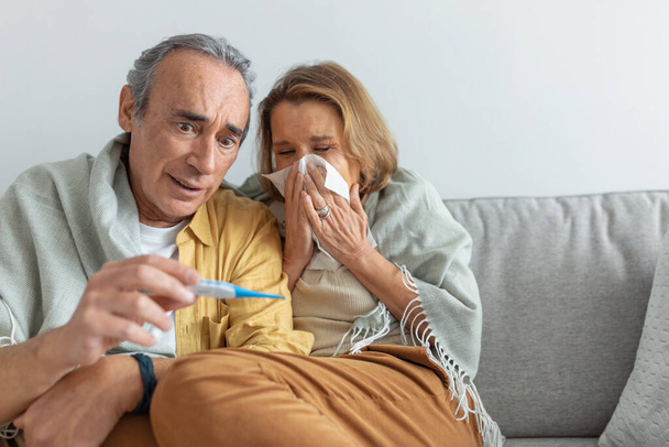 Shocked senior man measuring wifes temperature and looking at thermometer while woman sneezing in napkin, husband taking care of ill woman at home, copy space - Fotografie, Obrázek