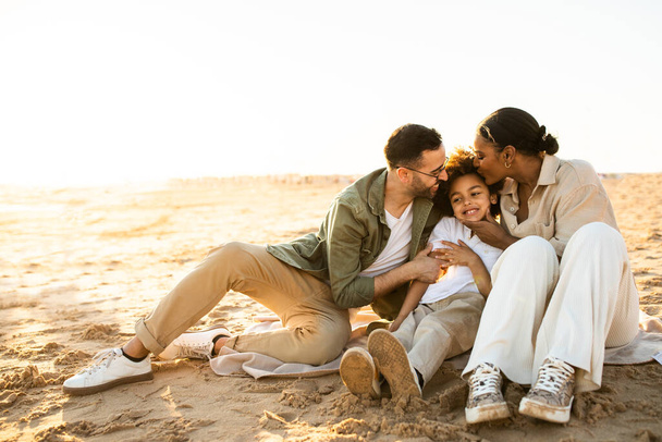 Щасливі багаторасові батьки обіймають і цілують сина, сидячи і відпочиваючи на пляжі біля моря, насолоджуючись ніжними моментами разом на заході сонця
 - Фото, зображення