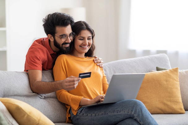 ラップトップと自宅でクレジットカードを介して幸せなインドのカップルのショッピング、若い東の配偶者は自宅でソファの上にリビングルームで座り、支払いを行い、コンピュータ上でインターネットバンキングサービスを使用して、スペースをコピーします - 写真・画像
