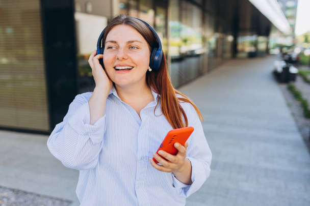 Egy boldog üzletasszony sétál ki a szabadba, és fülhallgatón keresztül telefonál. Elfoglalt nő telefonnal és fülhallgatóval az iroda közelében. Technológiát alkalmazó szakmai menedzser - Fotó, kép