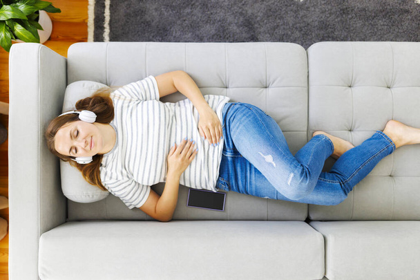 Peaceful woman in modern wireless headphones laying, relaxing on a comfortable couch, listening to music. Happy calm woman wearing headphones relaxing on a comfortable sofa, enjoy quality sound - Photo, image