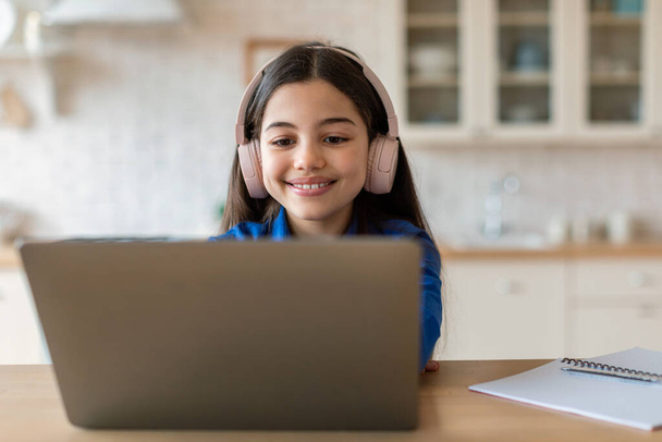 Az e-tanulás öröme. Happy Schoolgirl Böngészés Internet Laptop viselő fülhallgató, Online tanulás a számítógép ül otthon. Oktatás és kütyü. Élvezze a távoktatás koncepcióját - Fotó, kép