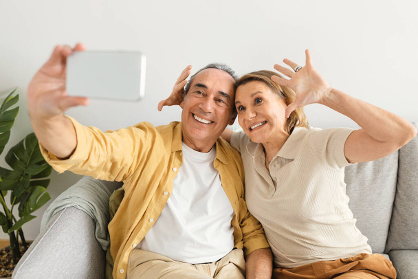 Χαρούμενο αστείο ζευγάρι ηλικιωμένων που παίρνει selfie στο smartphone, απολαμβάνοντας το χρόνο μαζί στο σπίτι ή έχοντας βιντεοκλήση με την οικογένεια. Αγάπη, ρομαντισμός, μοντέρνοι παππούδες - Φωτογραφία, εικόνα