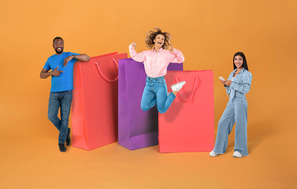 Consumerism, retail, online shopping, season sale, black friday, ecommerce. Cheerful multicultural millennials posing by huge colorful shopping bags over orange background, using smartphone, collage - Photo, Image