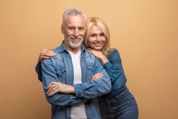 Studio portrait of casual beautiful elderly couple friends posing on beige background, happy senior handsome grey-haired bearded man and pretty blonde woman embracing, smiling at camera - 写真・画像
