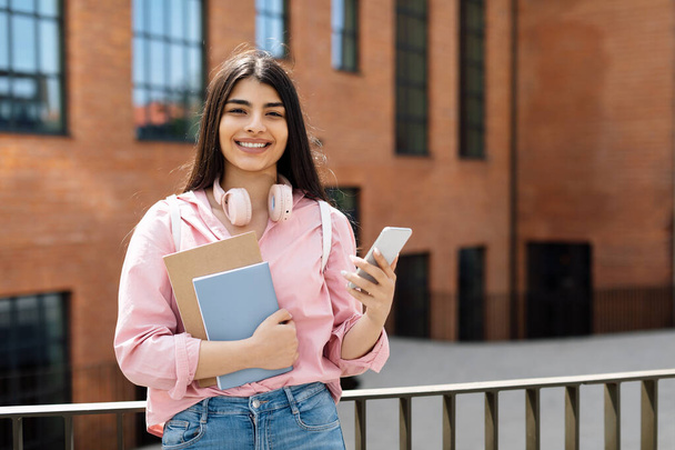 Portrait de dame étudiante latine joyeuse avec smartphone et classeurs posant à l'extérieur et souriant à la caméra, femelle utilisant le téléphone tout en se tenant à l'extérieur près de l'université - Photo, image