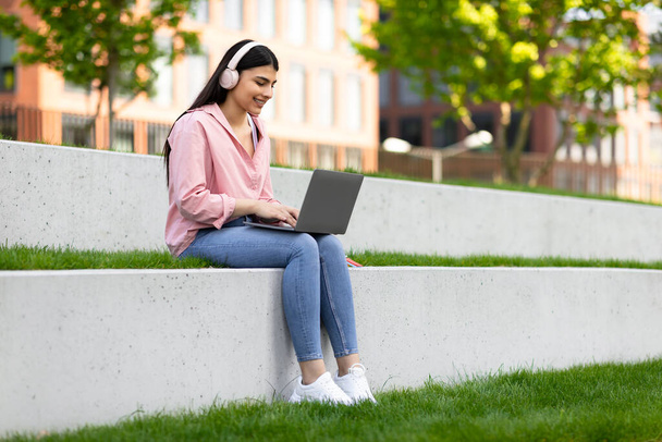 Smiling hispanic female student in wireless headphones studying with laptop outdoors, sitting in college campus or park, copy space. Happy lady enjoying distance learning - Foto, Imagem