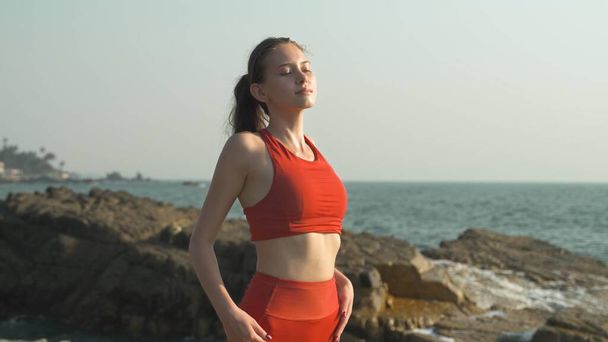 giovane donna in una tuta sportiva rossa con gli occhi chiusi si erge su uno sfondo di rocce e l'oceano. Foto di alta qualità - Foto, immagini