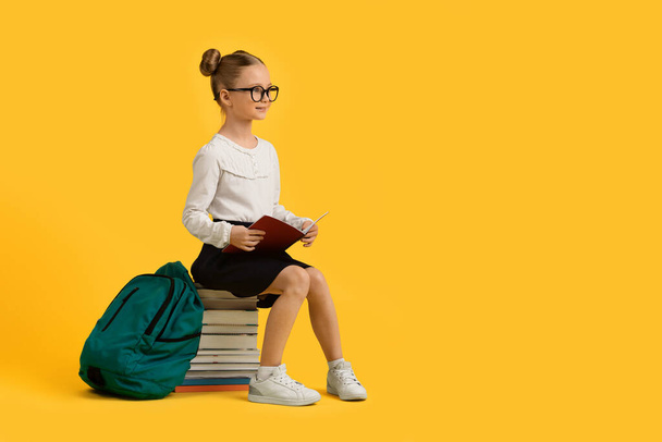 Studienkonzept. Lächelndes kleines Schulmädchen, das auf einem Stapel Bücher sitzt und den Kopierraum vor gelbem Studiohintergrund betrachtet, süßes, junges Mädchen, das eine Brille trägt und Arbeitsbuch in der Hand hält - Foto, Bild