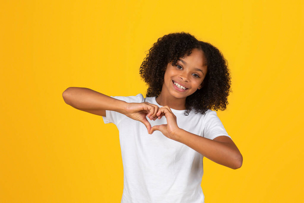 Positive curly teen black schoolgirl in white t-shirt has fun, making heart gesture with hands, isolated on yellow background, studio. Lifestyle, study, knowledge, gratitude and love sign, offer - Foto, afbeelding