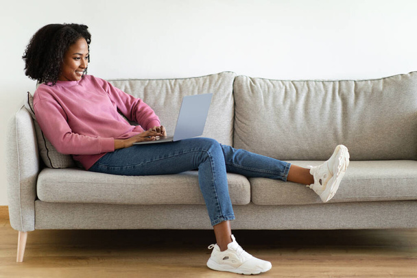 Pozytywne millenium czarna kobieta czatuje na komputerze, siedzi na kanapie we wnętrzu salonu, pełna długość. Praca, nauka, czat w sieciach społecznościowych, odpoczynek w domu, rozmowy wideo - Zdjęcie, obraz