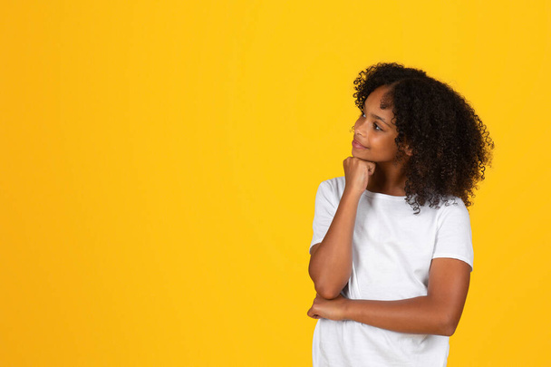 Happy pensive krullend tiener zwart schoolmeisje in wit t-shirt denkt, kiest, kijkt naar lege ruimte, geïsoleerd op gele achtergrond, studio. Studie vragen, kennis, reclame en aanbod - Foto, afbeelding