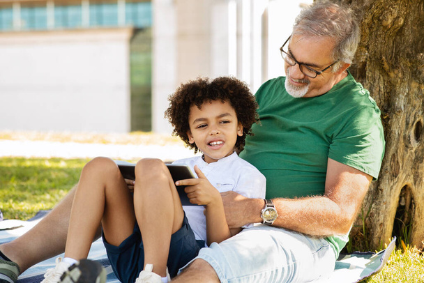 Cheerful senior man with beard and black little boy hugging, have fun, relax in park, watch video on tablet, outdoor. Relationship, grandson and grandfather, free time and walk, chat, game - Photo, Image