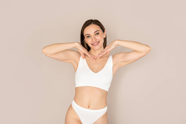Face care routine. Beatiful smiling young caucasian woman wearing white comfy underwear holding hands next to face, showing her beautiful smooth good-looking glowing skin, beige studio background - Valokuva, kuva