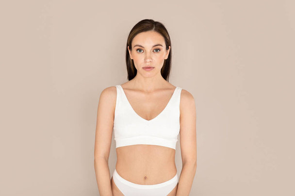 Body care concept. Attractive well-fit young caucasian woman posing in comfy white underwear on beige color background, showing nice body and silky youth-looking skin, copy space - Foto, Imagem