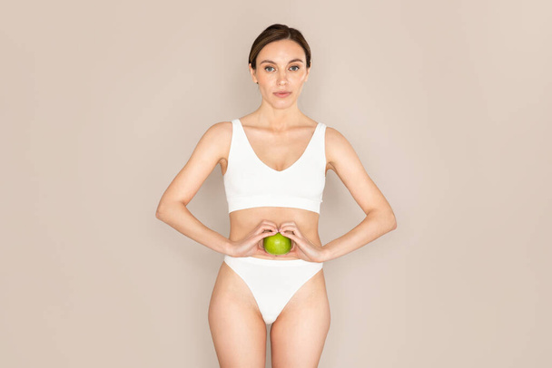 白い快適な下着を着た美しいスリムフィットの若い女性は、彼女の陰部腹、ベージュのスタジオの背景の横に緑のリンゴを保持。健康的な栄養、デトックス、食事、消化の概念 - 写真・画像